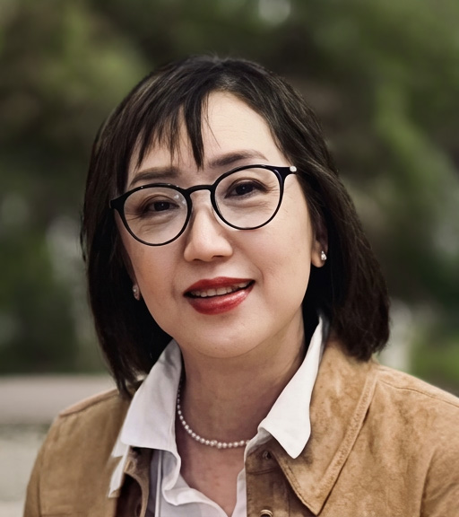 Portrait of a smiling woman with short black hair, round glasses, a brown jacket, white shirt, and a pearl necklace, outdoors with a blurred background.
